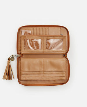 The Rip Curl Surf Gypsy Oversized tan leather wallet is open to reveal several compartments, including card slots, a zippered pocket, and transparent ID windows. An optional removable tassel keyring is attached to the zipper pull on the left side of the wallet. The background is white.