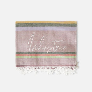 The Industrie The Maurepas Turkish Towel features a multicolored design with horizontal stripes in pink, yellow, green, and purple hues and "Industrie" written in white script across the center. Made of soft cotton, it is finished with elegant white fringes at the bottom edge.
