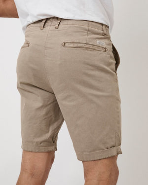 A person wearing the Industrie The Rinse Shorts in light brown, featuring a rolled hem and comfortable casual fabric, stands facing away. The shorts have two back pockets with visible stitching and are paired with a white t-shirt.
