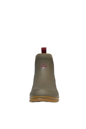 The Muck Boot Originals Women's Ankle Boot is shown from the front, showcasing its olive green rubber with a brown sole and textured surface. A red pull tab and prominent red diamond logo complete this waterproof shoe with an antimicrobial footbed, all set against a crisp white background.