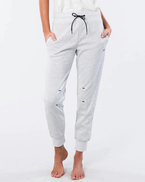 A person is standing barefoot against a plain background, wearing the Rip Curl Anti Series Flux II Trackpant in light gray, featuring a black drawstring. Their hands are in the pockets of the relaxed-fit pants, which also display small black details. They are paired with a white top.