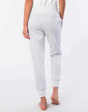 A person is standing barefoot on a light grey floor, facing away from the camera. They are wearing Rip Curl Anti Series Flux II Trackpants, renowned for their durable water-resistant fabric. Only the lower half of their body is visible against a plain white background.