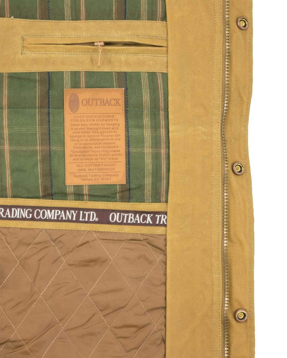 Close-up view of the interior of the Outback Gidley Jacket, showcasing its tan exterior with green plaid lining. The label reads "OUTBACK" alongside care instructions, and "Outback Trading Company LTD" is printed on an inner fabric band. This comfortable jacket features quilted padding for warmth and dual entry pockets for convenience.