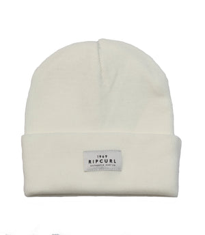 The Rip Curl Premium Surf Beanie is a white rib knit hat featuring a folded brim. A rectangular fabric patch on the front of the brim showcases the text "1969 RIP CURL A PRODUCT OF THE SURF CO." in bold black capital letters. This beanie boasts a simple, clean design.