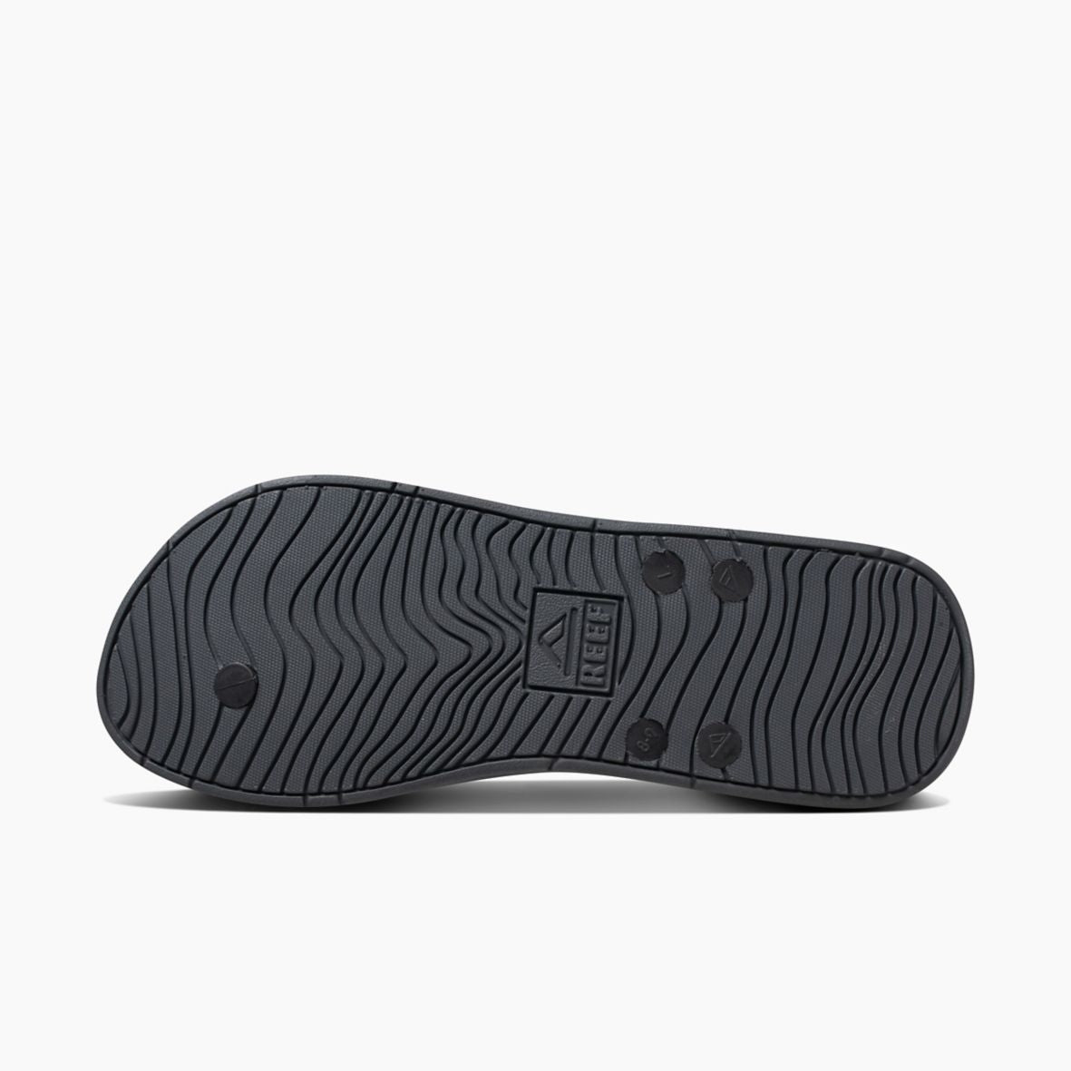 The image showcases the black rubber outsole of the REEF Switchfoot LX sandal, featuring the "REEF" logo imprinted in the center. The sole has wavy grooves for traction and small circular patterns at various points for added grip, while its rubber sponge footbed offers excellent arch support.