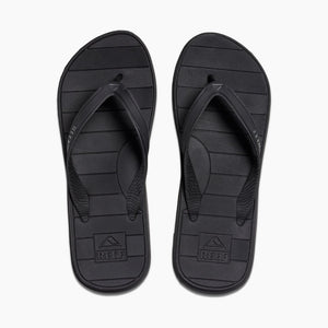 A pair of REEF Switchfoot LX flip-flops in black features a simple striped pattern on the rubber sponge footbed. The brand name "Reef" is embossed on the heel area of each sandal, with subtle text and logo detailing on the straps. These flip-flops, designed with arch support, are displayed against a plain white background.