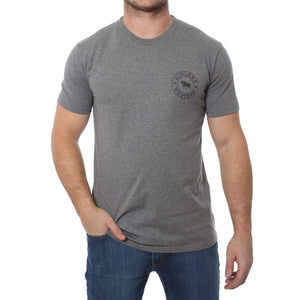 A person is standing with one hand in a pocket, wearing the Ringers Western Signature Bull Classic T-Shirt in plain grey, featuring a small round emblem with text and a graphic on the left chest area, paired with blue jeans. The face is not visible.