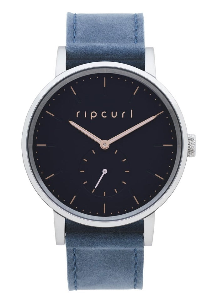 The Rip Curl Circa Leather Watch embodies style with its dark blue face and silver casing, meticulously crafted from marine-grade stainless steel. The face prominently displays the brand name "Rip Curl" in gold letters, complemented by gold hour markers and a small sub-dial at 6 o'clock. To complete its elegant design, the watch features a genuine leather band in matching blue.