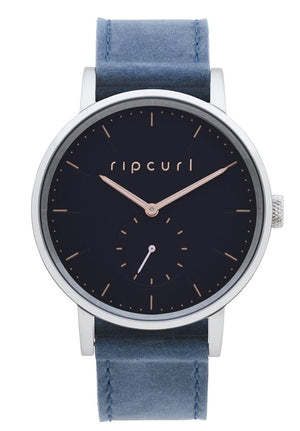 The Rip Curl Circa Leather Watch embodies style with its dark blue face and silver casing, meticulously crafted from marine-grade stainless steel. The face prominently displays the brand name "Rip Curl" in gold letters, complemented by gold hour markers and a small sub-dial at 6 o'clock. To complete its elegant design, the watch features a genuine leather band in matching blue.