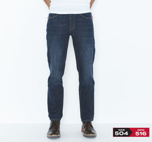 A person wearing dark blue Levis 516 Straight Jeans and brown shoes stands against a plain white background. The left corner of the image shows a red and black price tag indicating that the 100% cotton jeans were previously priced at 504 and are now priced at 516.