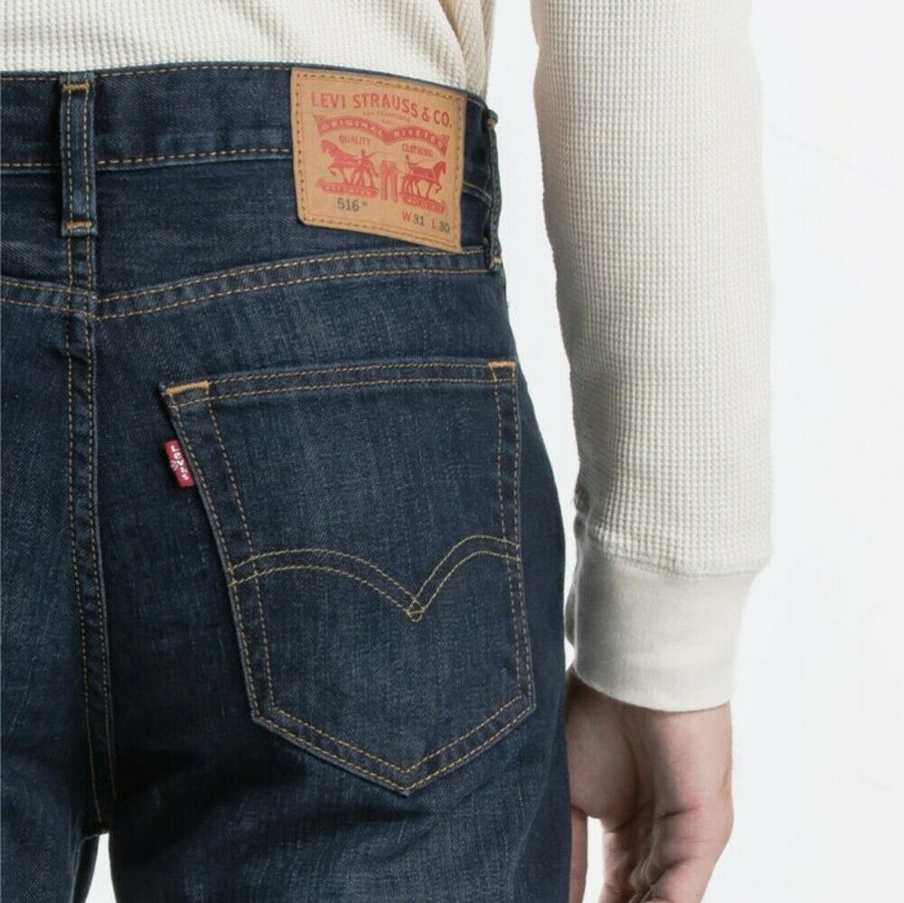 A close-up view of a person wearing dark blue Levis 516 Straight Jean, highlighting the back pocket and the brown patch on the waistband featuring the Levi Strauss & Co. logo. The 100% cotton jeans are paired with a cream-colored long-sleeve shirt for a comfortable fit.