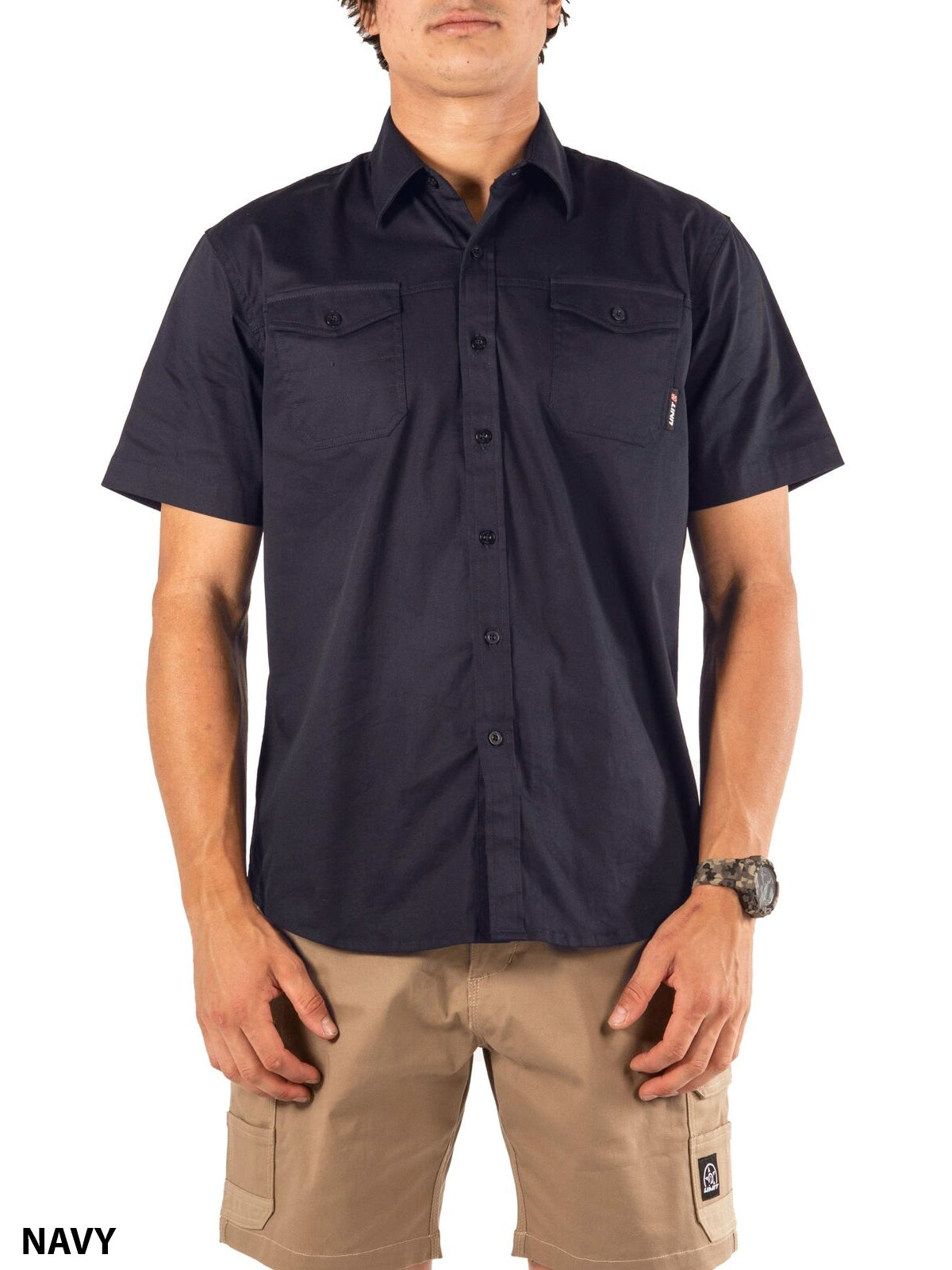 A person wears the UNIT Craftman Stretch Short Sleeve Shirt in navy with two chest pockets and tan cargo shorts, facing forward. "NAVY" is displayed in the bottom left corner.
