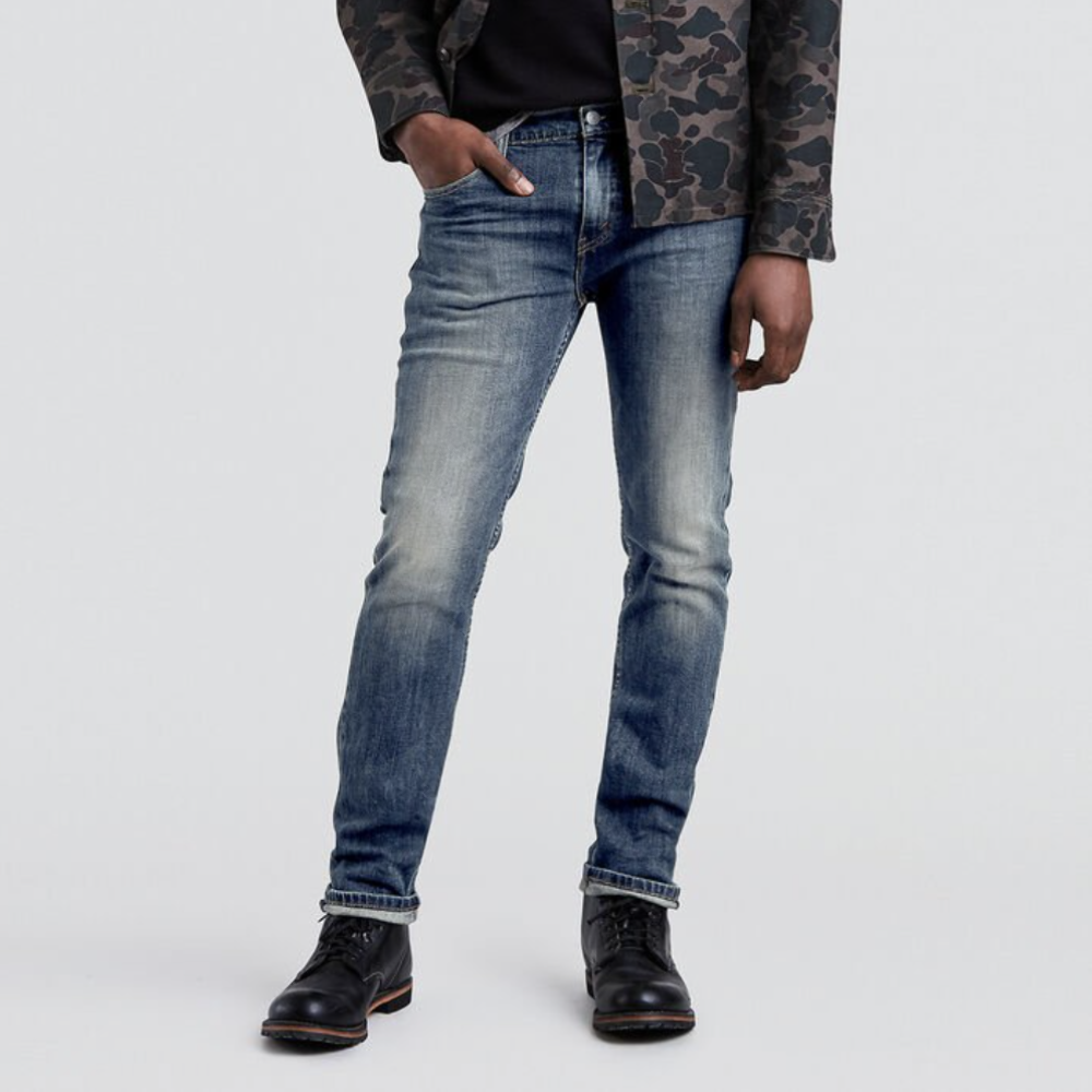 A person is standing against a plain white background, wearing a camouflage jacket, dark shirt, and black boots. The focus is on the lower body, highlighting their faded Levi's 511 Slim Jean with hands resting casually in their pockets.