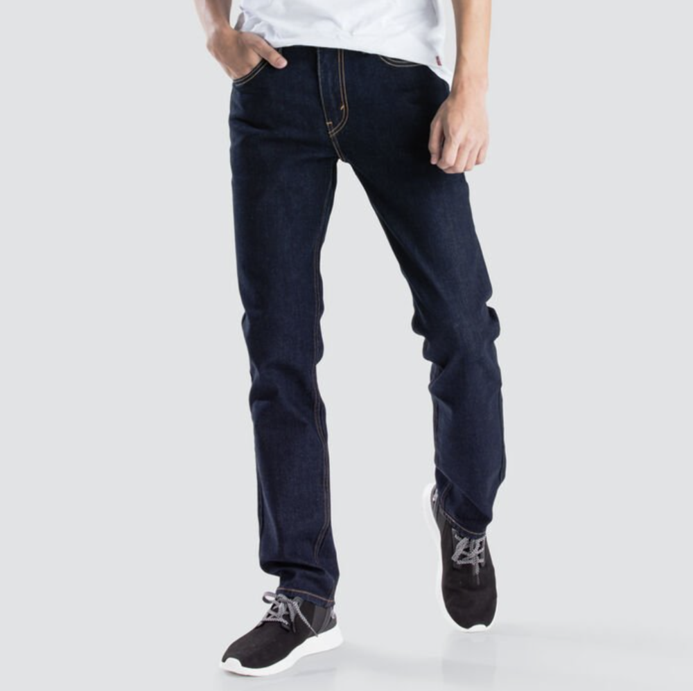 A person wearing stylish and comfortable dark blue Levis 511 Slim Jeans, a white shirt, and black sneakers poses against a plain light gray background. The individual's left hand is in their slim fit jeans pocket, while the right arm hangs by their side. The head is not visible in the frame.