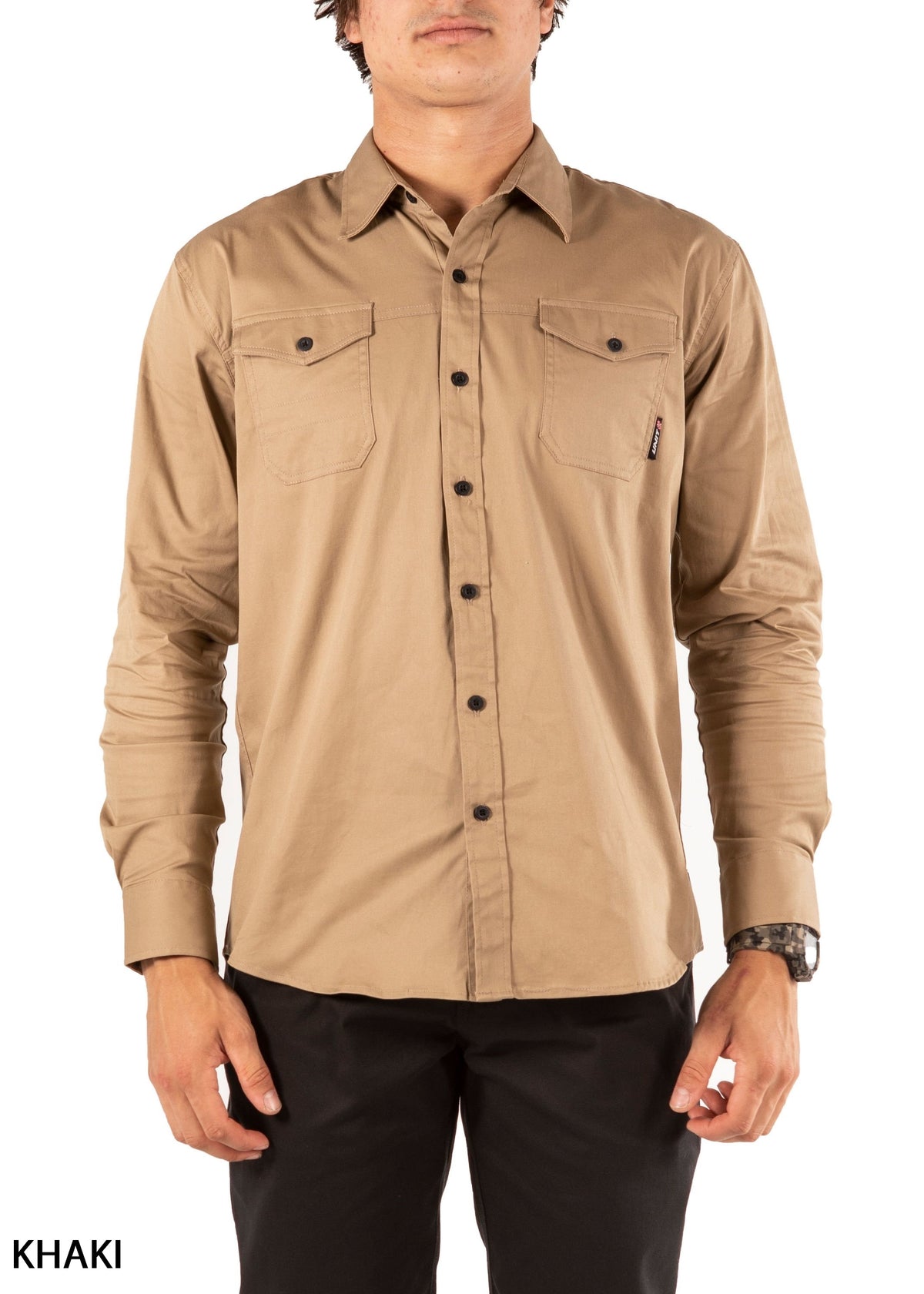 A person wears the UNIT Craftman Stretch Shirt—a long-sleeve, button-up khaki work shirt with cotton stretch and two chest pockets—paired with black pants, standing before a white background. "KHAKI" appears in the bottom left.