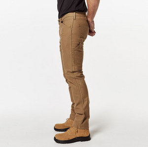 Side view of a person standing while wearing comfortable light brown Levis Workwear 511 Utility Pants, a black shirt, and tan work boots. The individual’s hands are clasped behind their back. The image is set against a plain white background.