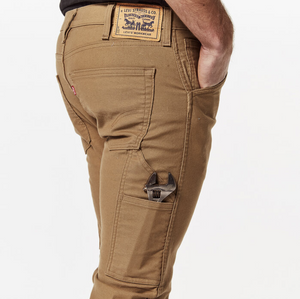 A close-up image features a person wearing Levi's Workwear 511 Utility Pant in caramel color, highlighting intricate stitching details. An adjustable wrench is neatly tucked into the side utility pocket, and the Levi's brand patch is prominently displayed on the back waistband. The individual, comfortably posed with their hand in the pocket, exemplifies a blend of sleek style and practical functionality.