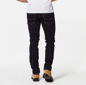 A person stands with their back facing the camera, comfortably dressed in Levis Workwear 511 Slim Jean in dark blue and a white t-shirt. They wear light brown boots, with their hands resting by their sides against a plain white background.