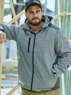 A man wearing a Bisley Flx & Move Shield Jacket in gray, paired with yellow pants and a dark cap that says "Bisley," is standing outdoors at a construction site. He has a contemplative look and is leaning against a wooden structure.