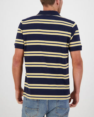 A person wearing a Swanndri Lincoln Stripe Polo, featuring navy blue with yellow and white horizontal stripes, and blue jeans is shown from the back. The 100% cotton casual polo shirt complements their short hair as they stand against a plain, light-colored background.