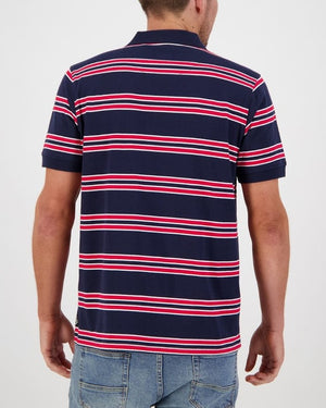 A person with short hair is seen from the back wearing the Swanndri Lincoln Stripe Polo, featuring casual navy blue fabric with red and white horizontal stripes made of 100% cotton. They are also wearing blue jeans against a plain white background.