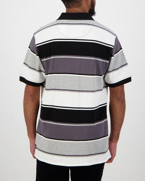 A man with short hair and a beard faces away from the camera, dressed in a Swanndri Kingfield Stripe Polo. This 100% cotton shirt boasts black, gray, and white horizontal stripes with a classic black collar, exuding casual elegance.