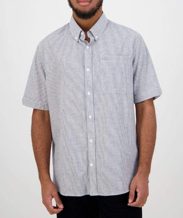 In front of a plain backdrop, someone wears the Swanndri Hinsdale Shirt, a classic fit, short-sleeved button-up with thin vertical stripes and a chest pocket. Their hands rest at their sides. Made from 100% cotton.