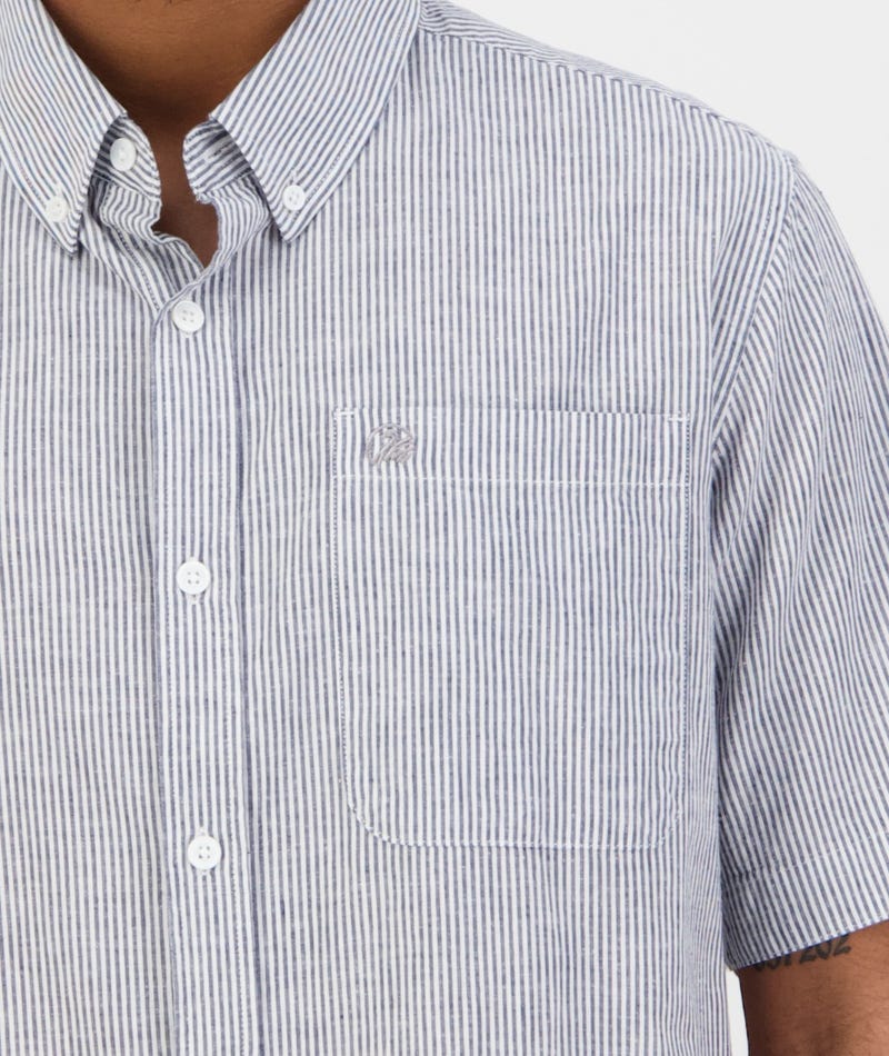 A person wears the Swanndri Hinsdale Shirt, a classic-fit blue and white striped button-up with a left-side pocket and small embroidered logo above it. Made from 100% cotton, the comfortable shirt reveals the person's arm and part of their neck.