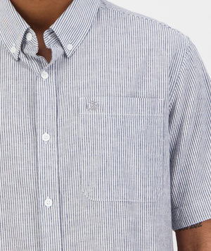A person wears the Swanndri Hinsdale Shirt, a classic-fit blue and white striped button-up with a left-side pocket and small embroidered logo above it. Made from 100% cotton, the comfortable shirt reveals the person's arm and part of their neck.
