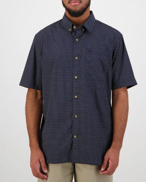 A person models against a plain backdrop in the Swanndri Putney Shirt, a tailored-fit navy short-sleeve button-up featuring a subtle white pattern. This 100% cotton shirt with gold buttons complements their beige pants and beard effortlessly.