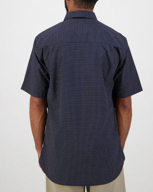 The individual is shown from behind wearing a tailored-fit Swanndri Putney Shirt, a dark short-sleeve shirt with small white polka dots made from 100% cotton. Paired with beige pants, this ensemble stands out against the plain, neutral background.
