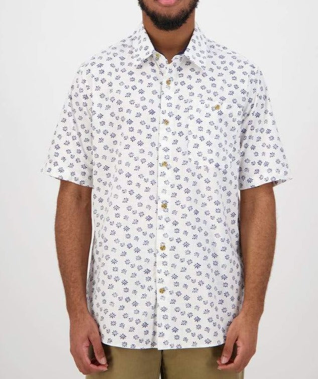 A man wearing a comfortable Swanndri Belfast Shirt, a short-sleeved white button-up featuring a blue floral pattern, stands facing forward. He has a beard and is dressed in light brown pants. This casual shirt is made of 100% cotton, and the background is plain white.