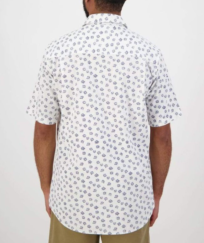 A person with short hair is seen from the back, wearing a comfortable, casual Swanndri Belfast Shirt, which is a white short-sleeved button-up with a blue floral pattern, paired with beige pants. The background is plain white.