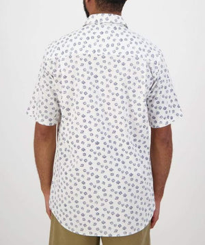 A person with short hair is seen from the back, wearing a comfortable, casual Swanndri Belfast Shirt, which is a white short-sleeved button-up with a blue floral pattern, paired with beige pants. The background is plain white.