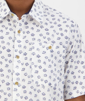 Close-up of a person wearing the Swanndri Belfast Shirt, a comfortable, casual shirt crafted from 100% cotton. This white short-sleeve button-up shirt features a striking blue floral pattern and includes a pocket on the left side secured with a button. The design is completed with a pointed collar and light brown buttons.