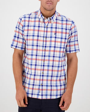A person is wearing the Swanndri Taunton Shirt, a classic fit, short-sleeved button-up featuring a checkered pattern in blue, red, and white colors. The comfortable shirt has a small embroidered logo on the chest. They are standing against a white background with their arms relaxed by their sides.