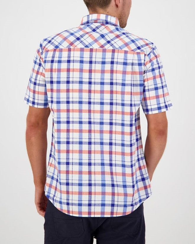 A man is standing with his back to the camera, dressed in a classic fit short-sleeved Swanndri Taunton Shirt featuring a red, white, and blue plaid pattern, paired with dark-colored pants. The setting is well-lit and the background is plain white.