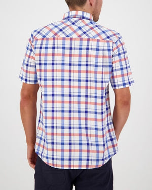 A man is standing with his back to the camera, dressed in a classic fit short-sleeved Swanndri Taunton Shirt featuring a red, white, and blue plaid pattern, paired with dark-colored pants. The setting is well-lit and the background is plain white.