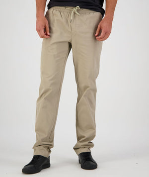 A person stands against a plain background, modeling Swanndri Karaka Pant in beige with an elastic waist and black sneakers. The white drawstrings are tied in a bow at the waist. The person is wearing a dark top that is partially visible in the frame.