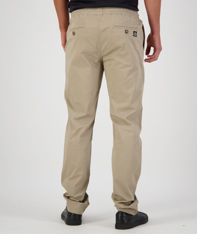 A person wearing beige Swanndri Karaka Pants with buttoned back pockets, a black shirt, and black shoes is standing facing away from the camera. The pants feature an elastic waist for added comfort. The person is positioned against a plain white background.