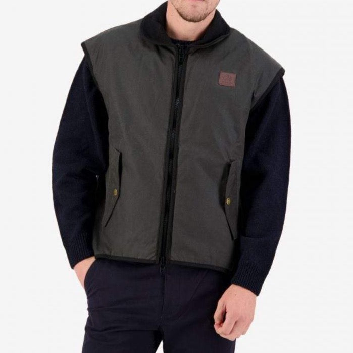 A man wears a Swanndri Foxton Vest with a dark green, waxed cotton, waterproof outer over a black sweater and dark pants, standing against a plain white background with one hand in his pocket.
