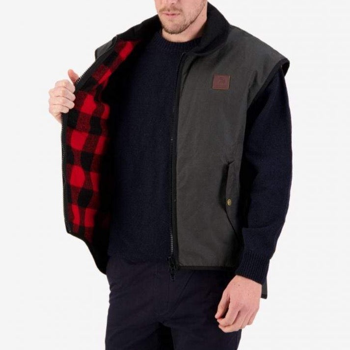 A person wears a Swanndri Foxton Vest Wool Lined, crafted from waxed cotton with a red and black plaid wool lining, over a navy sweater. The vest is unzipped and partially open, showcasing the lining against a plain white background.