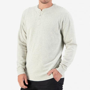 A person is shown from the waist up wearing a Swanndri Oxford Henley in light gray, featuring long sleeves and waffle knit detailing. The casual top includes a buttoned placket at the neckline. With one hand in their pocket, they are standing against a plain white background.