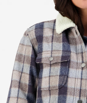 Close-up view of a person wearing the Swanndri Kaituna Women's Sherpa Lined Jacket, featuring a stylish blue and beige plaid design with a white sherpa collar and snap-button flap pockets on the chest. The person's head is partially visible, against a plain white background.