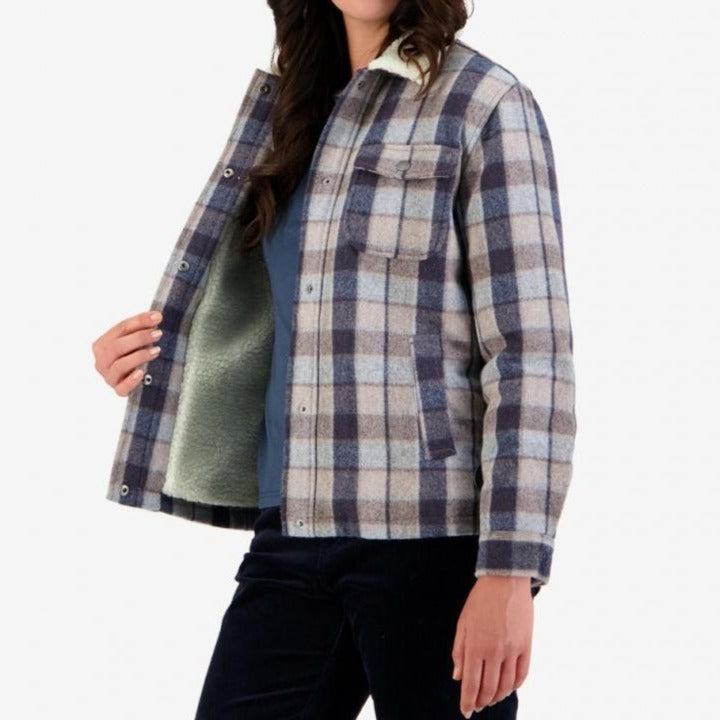 A person is modeling the Swanndri Kaituna Women's Sherpa Lined Jacket, a stylish blue and gray plaid jacket with snap buttons. The jacket, which features a cozy sherpa collar, is open to reveal a light green inner lining. The person is wearing a blue shirt and dark pants, with their face not visible.