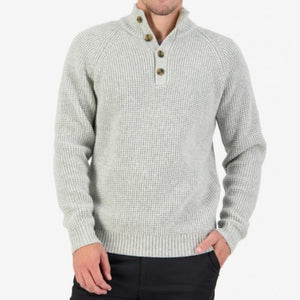 A person is wearing the Swanndri Okains Bay Button Neck Waffle Knit sweater in light grey, featuring a textured knit pattern with a high collar and three buttons at the neck. The individual has one hand in the pocket of their dark trousers, standing against a plain white background.