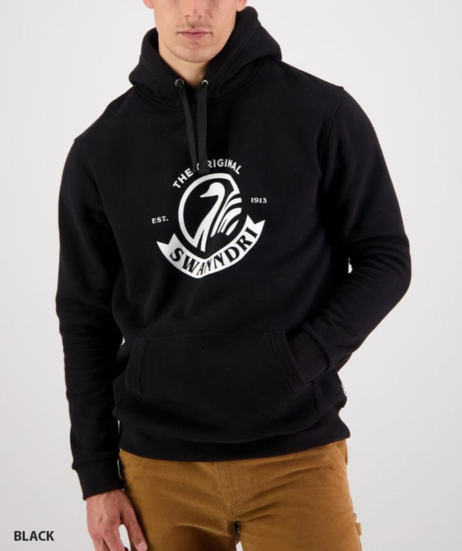A person wears the Swanndri Original Fleece Hoody, featuring a black design with a white "The Original Swanndri Est. 1913" logo. This casual hoodie, made of loop back fleece with front pockets, is stylishly paired with brown trousers.