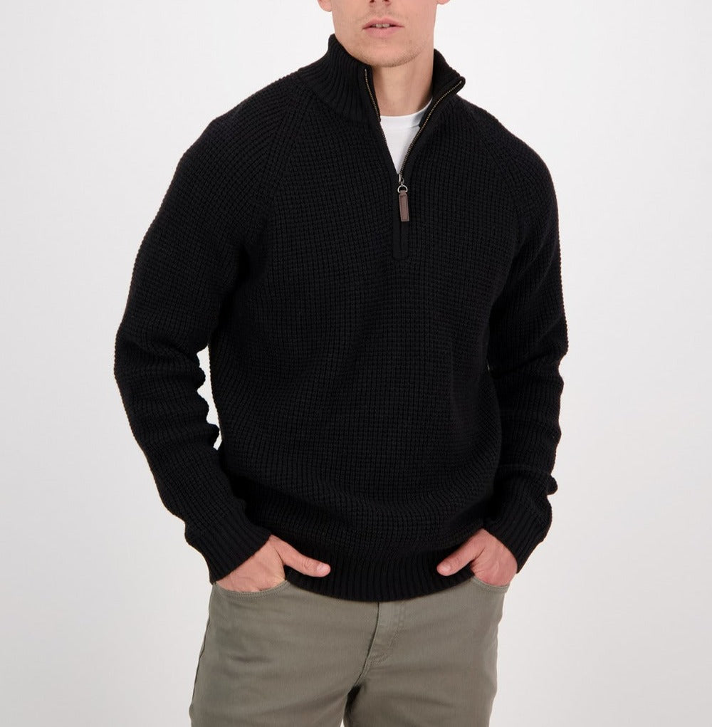 A man wearing the Swanndri Chalky Island Waffle Jumper, a black heavyweight knit half-zip sweater, over a white shirt and olive green pants stands with his hands in his pockets against a plain white background.