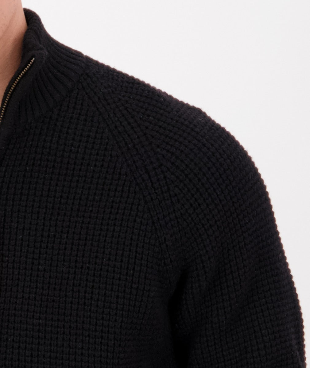 Close-up of a person wearing the Swanndri Chalky Island Waffle Jumper, a heavy knit black sweater with a zipper and ribbed collar, against a plain light background. Only part of the shoulder, chest, and neck is visible.