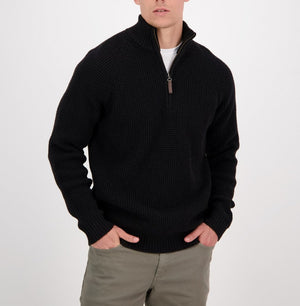 A man wearing the Swanndri Chalky Island Waffle Jumper, a black heavyweight knit half-zip sweater, over a white shirt and olive green pants stands with his hands in his pockets against a plain white background.